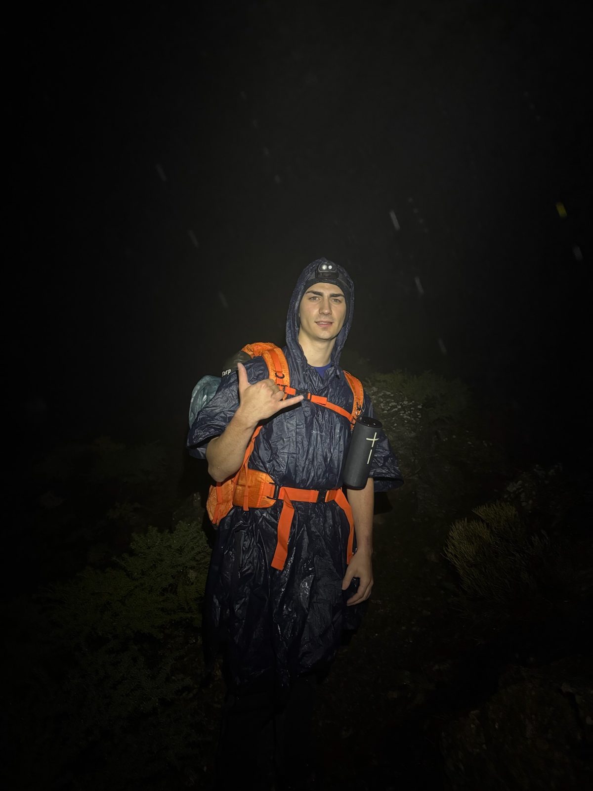 A hiker in a rain poncho and orange backpack, lit by a headlamp in the dark