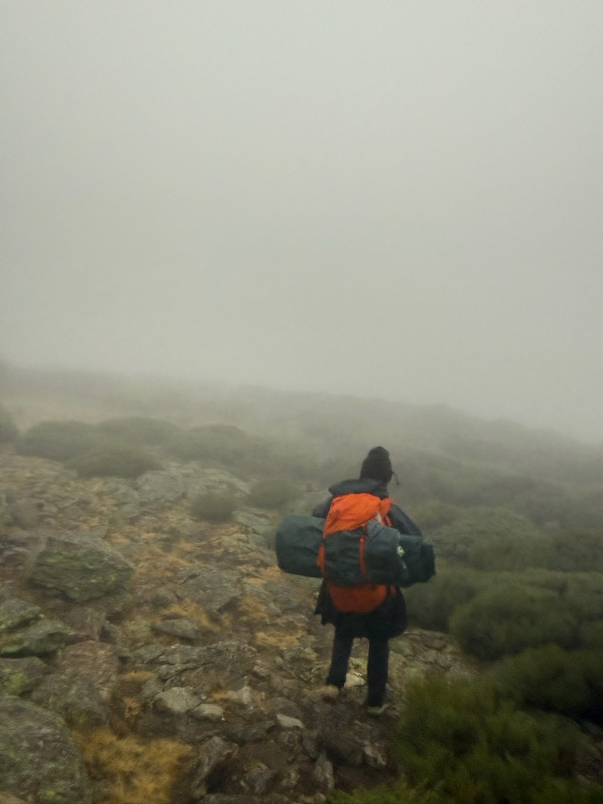 A hiker with an orange pack walking across rocky ground into fog